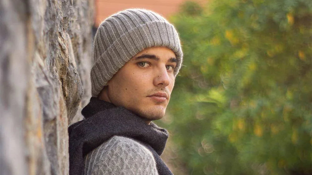 Man wearing a grey knit beanie and dark scarf, outdoors near stone wall, Norgäte luxury knitwear